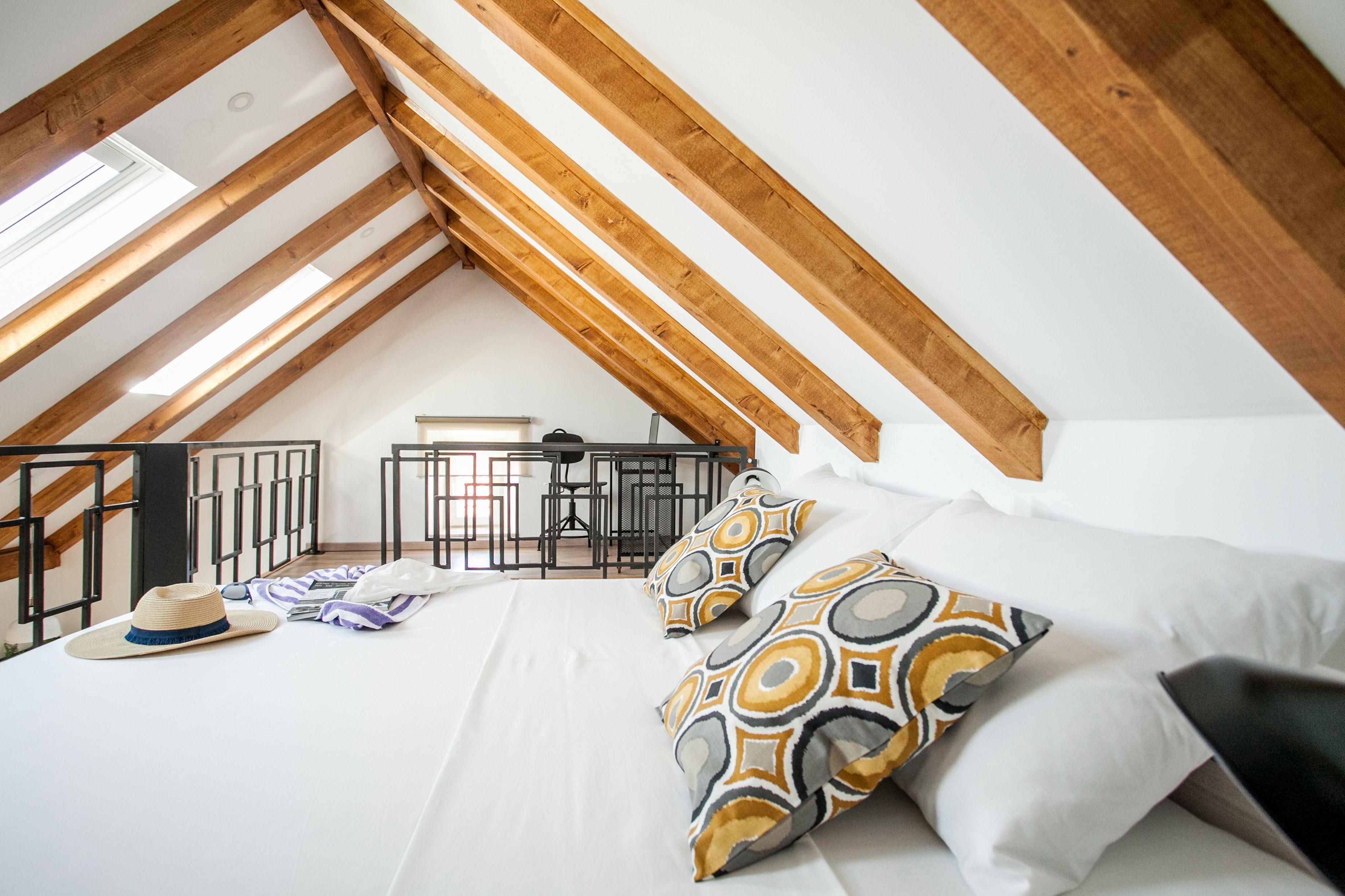 Un dormitorio con techo inclinado, una cama con almohadas estampadas, un sombrero y una maleta en el suelo.