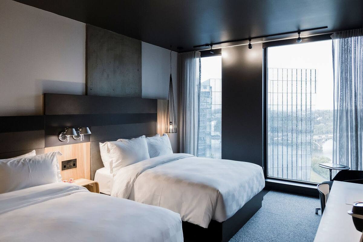 Quarto de hotel moderno com duas camas, uma janela grande com vista para a cidade e uma pequena mesa.