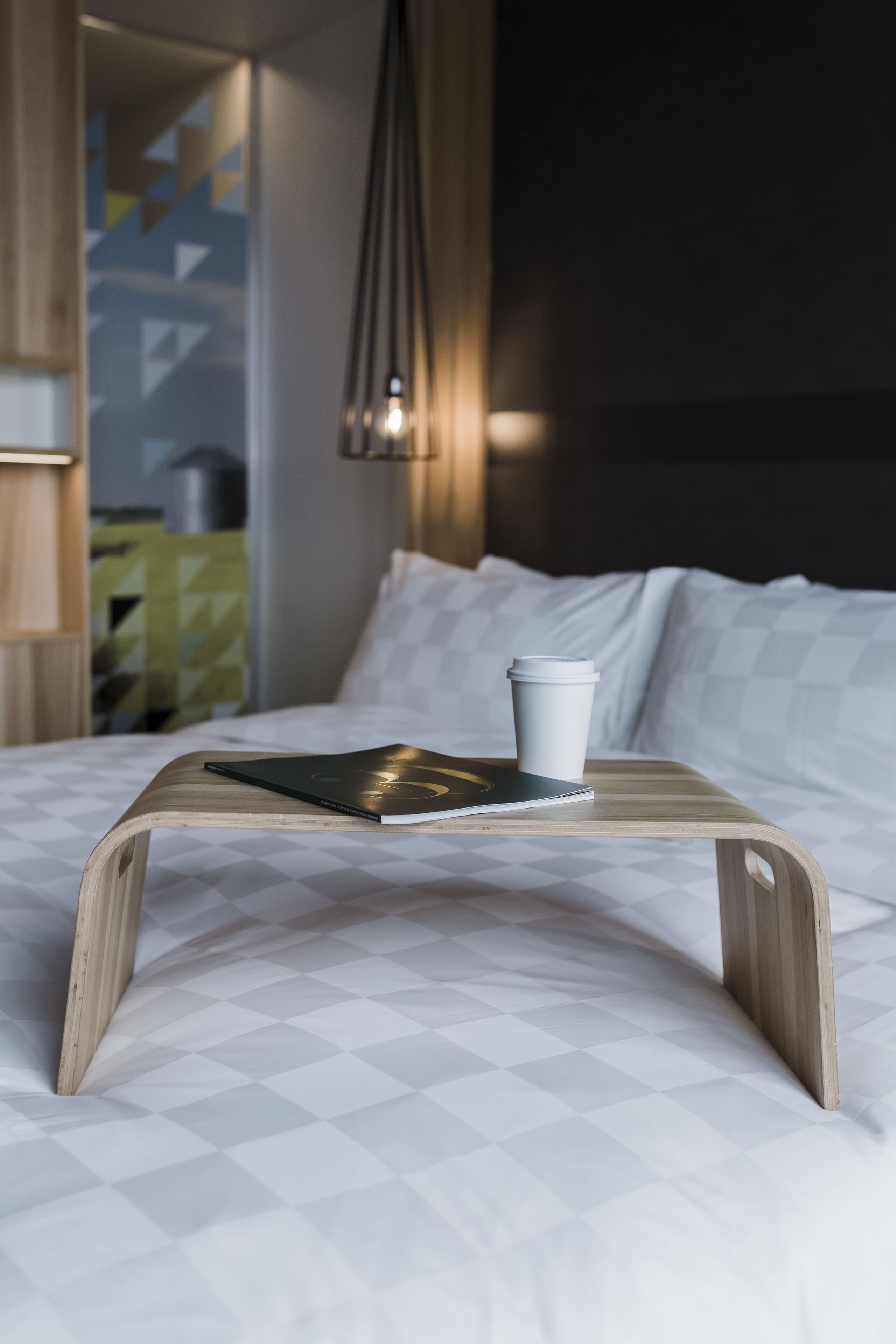 A neatly made bed with a white checkered bedspread, a wooden bedside table with a book and a white cup, and a modern pendant light hanging above.