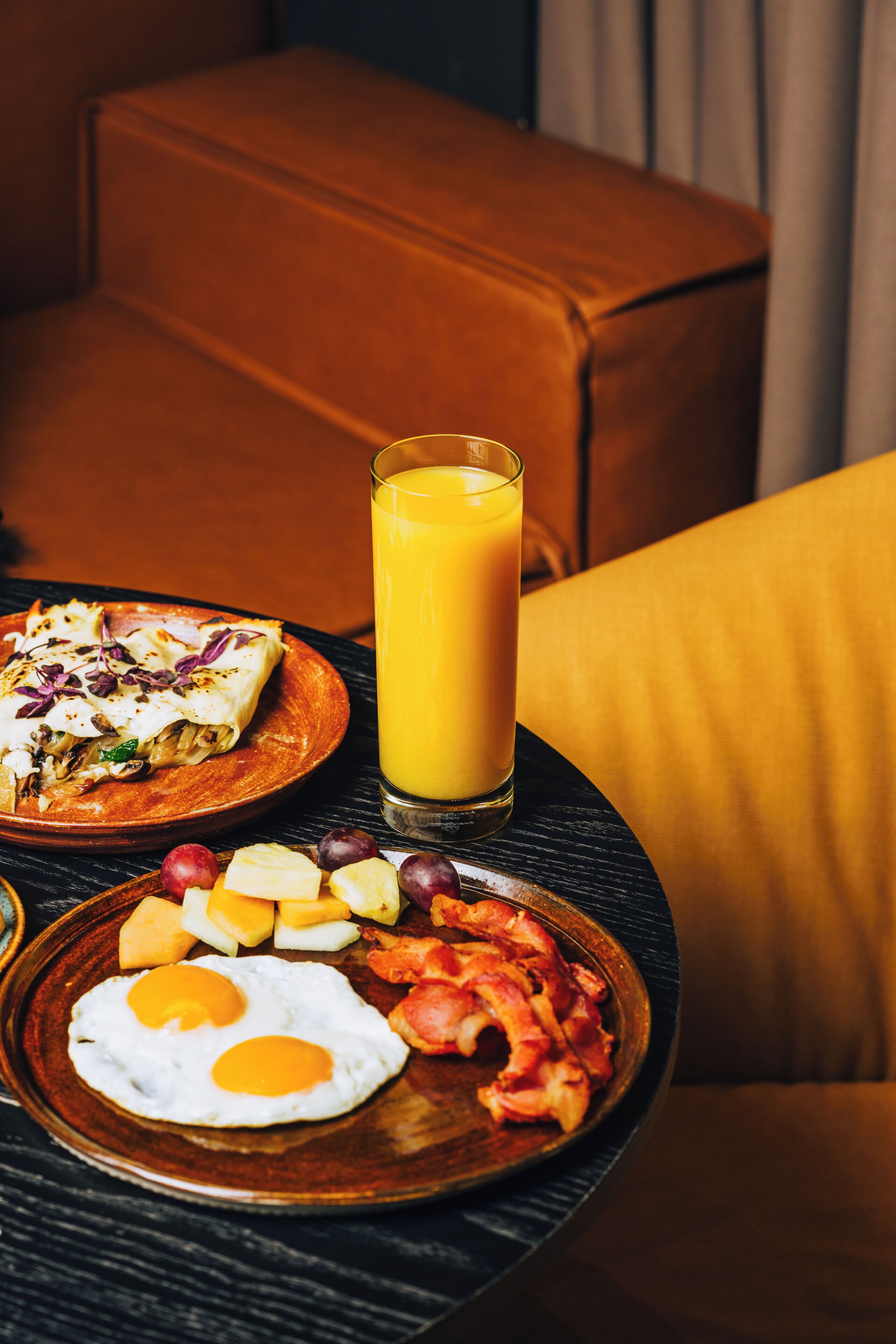 A breakfast setting with a plate of eggs, bacon, and fruit, a glass of orange juice, and a quesadilla on a table.