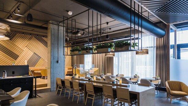 Un espacio de oficina moderno con una larga mesa compartida, varias sillas y una zona de cocina con plantas.