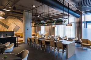 A modern office space with a long communal table, multiple chairs, and a kitchen area with plants.