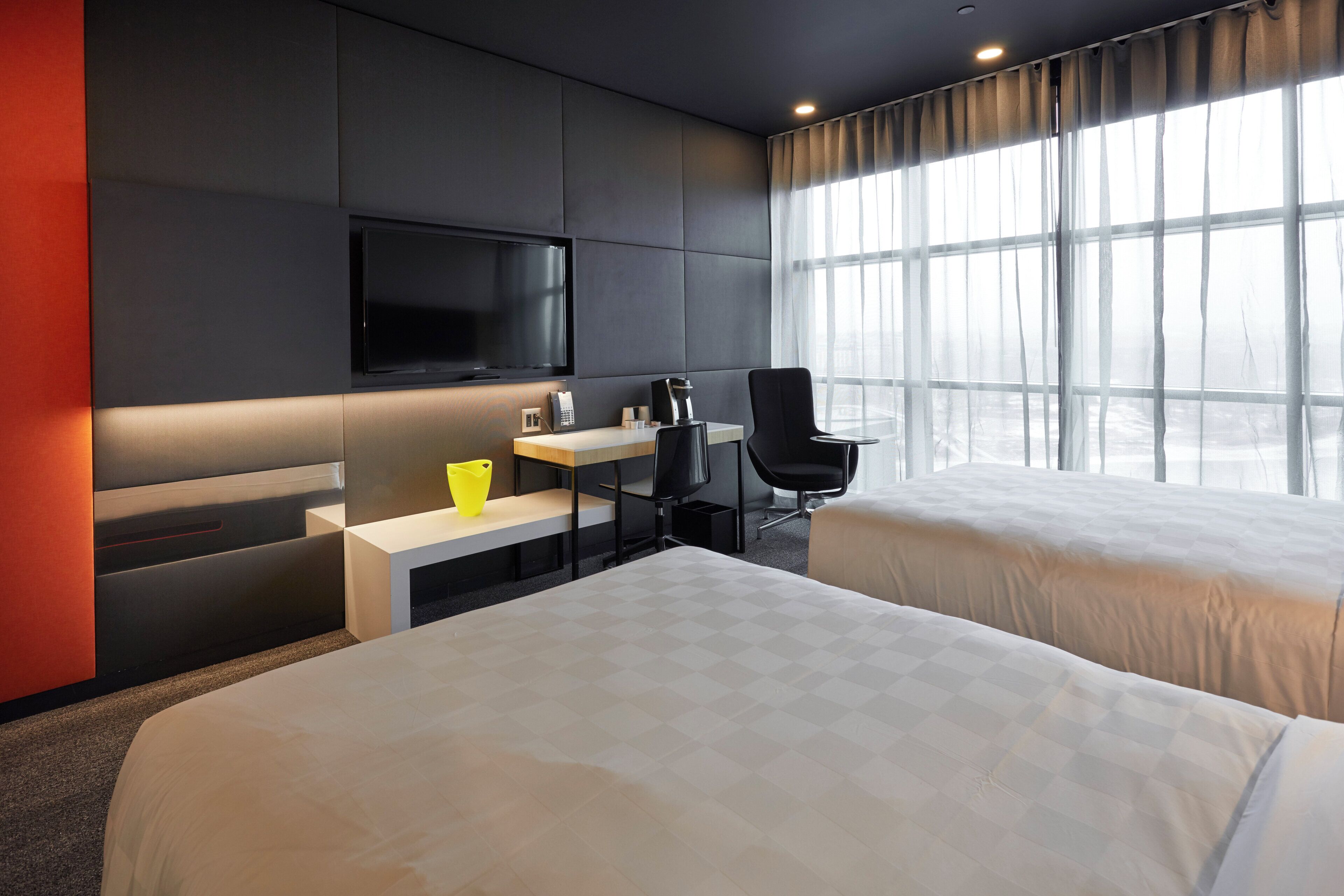 A modern hotel room with two beds, a desk with a coffee maker, a TV, and a large window with curtains.