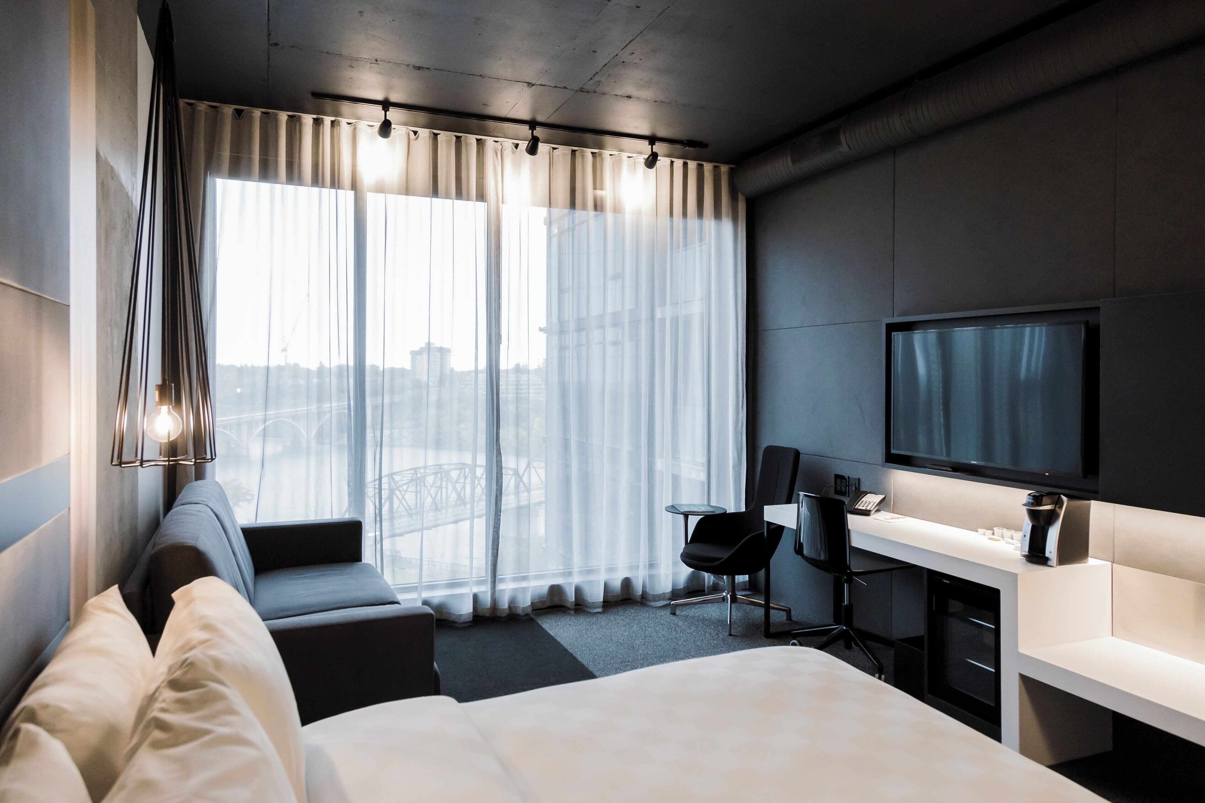 Una habitación de hotel moderna con una cama grande, un escritorio con televisor de pantalla plana, una silla y una ventana con cortinas translúcidas.