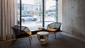 Una habitación moderna con amplios ventanales, dos sillas de malla negra con cojines amarillos, una mesa redonda de vidrio y una pared de concreto.