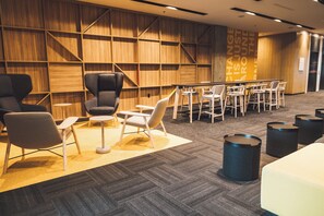A modern lounge area with a wooden wall, multiple seating options including chairs and tables, and a carpeted floor.