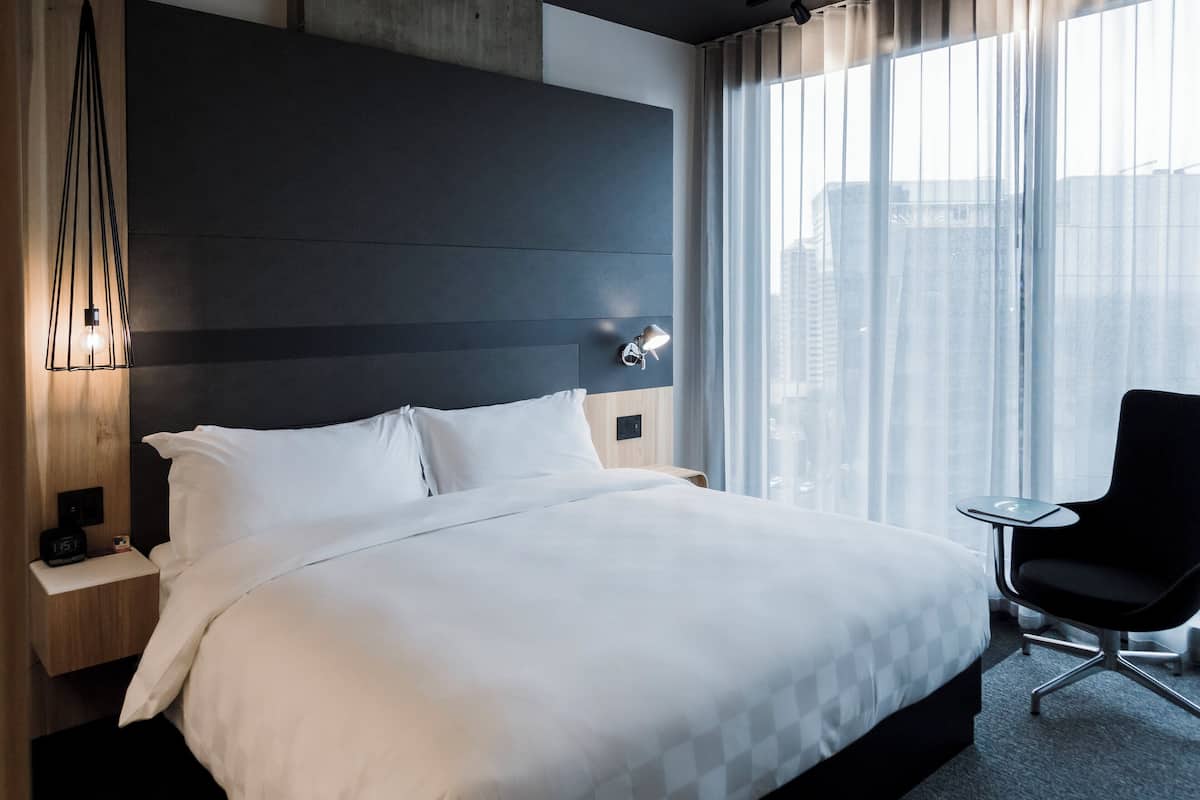Quarto de hotel moderno com uma cama grande, mesa de cabeceira com abajur, uma cadeira e vista da cidade através de cortinas translúcidas.