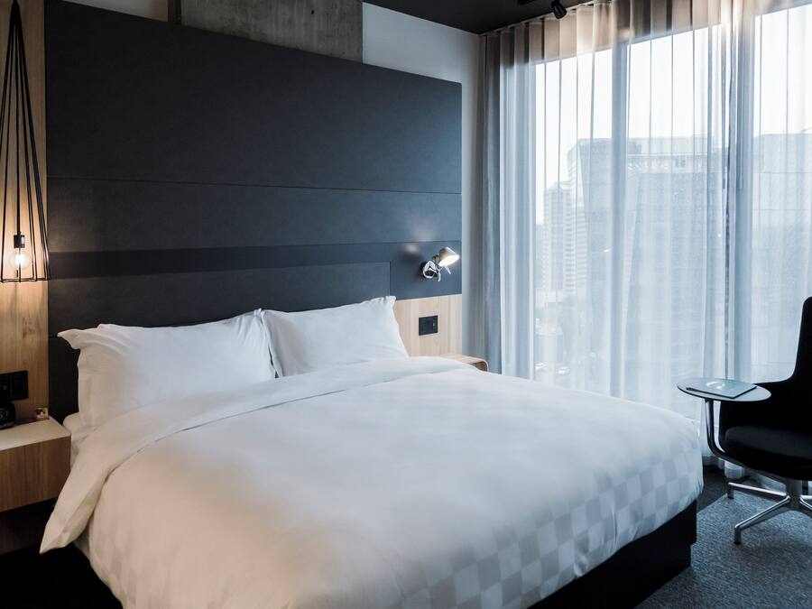 Un hotel moderno con una cama grande, una mesita de noche con lámpara, una silla y vistas a la ciudad a través de cortinas translúcidas.