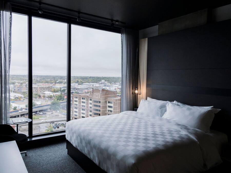 Habitación de hotel con un ventanal amplio que ofrece vistas a la ciudad, una cama bien hecha y una mesita pequeña.