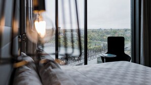 A hotel room with a bed, a chair, and a view of a cityscape through large windows. - Alt Hotel Saskatoon (Saskatoon)