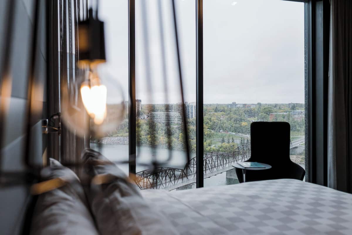 Quarto de hotel com uma cama, uma cadeira e vista para a cidade através de grandes janelas.