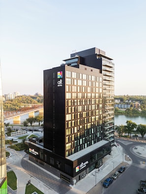 A modern hotel building with a prominent logo, a glass facade, and a view of a river and bridge in the background. - Alt Hotel Saskatoon (Saskatoon)