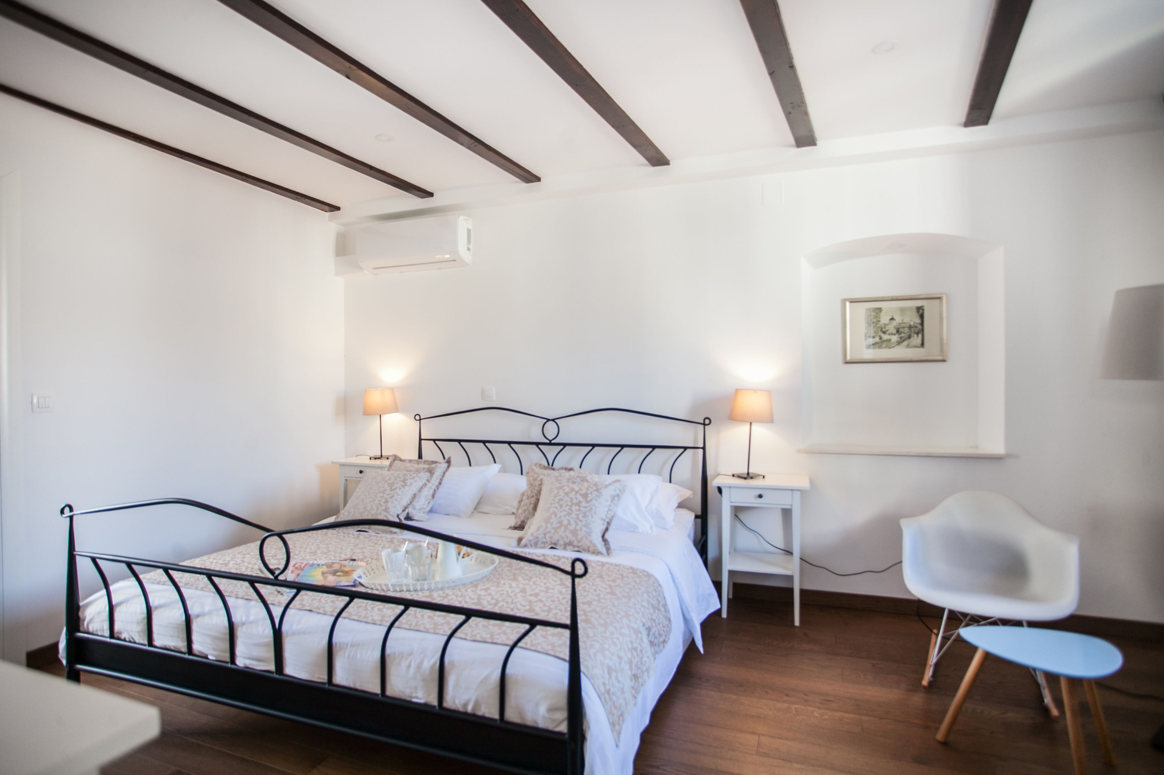 A bedroom with a metal bed frame, white walls, a wooden ceiling, a bedside table with a lamp, a chair, and a framed picture on the wall.