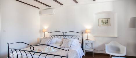 A bedroom with a metal bed frame, white walls, a wooden ceiling, a bedside table with a lamp, a chair, and a framed picture on the wall.
