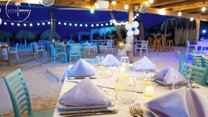 En restaurant ved stranden med borde dækket til spisning, lyskæder over hovedet og udsigt til havet.