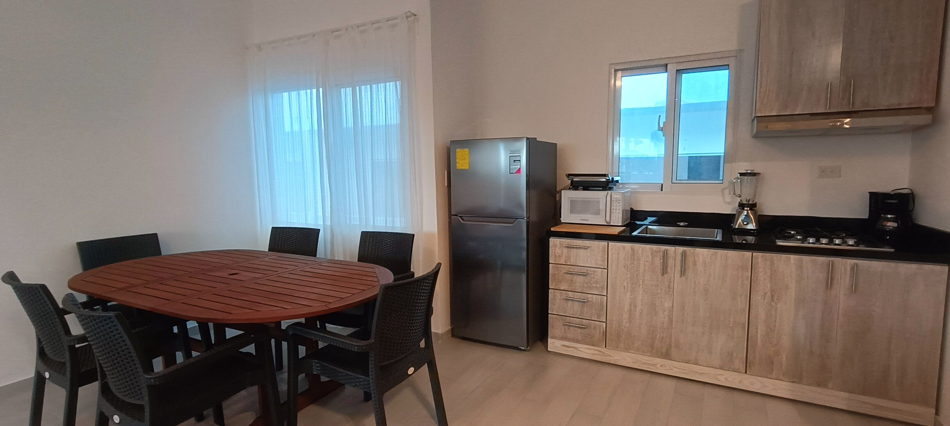 Cocina compacta con refrigerador, microondas y estufa. Hay una mesa de comedor con sillas y una ventana con cortinas.