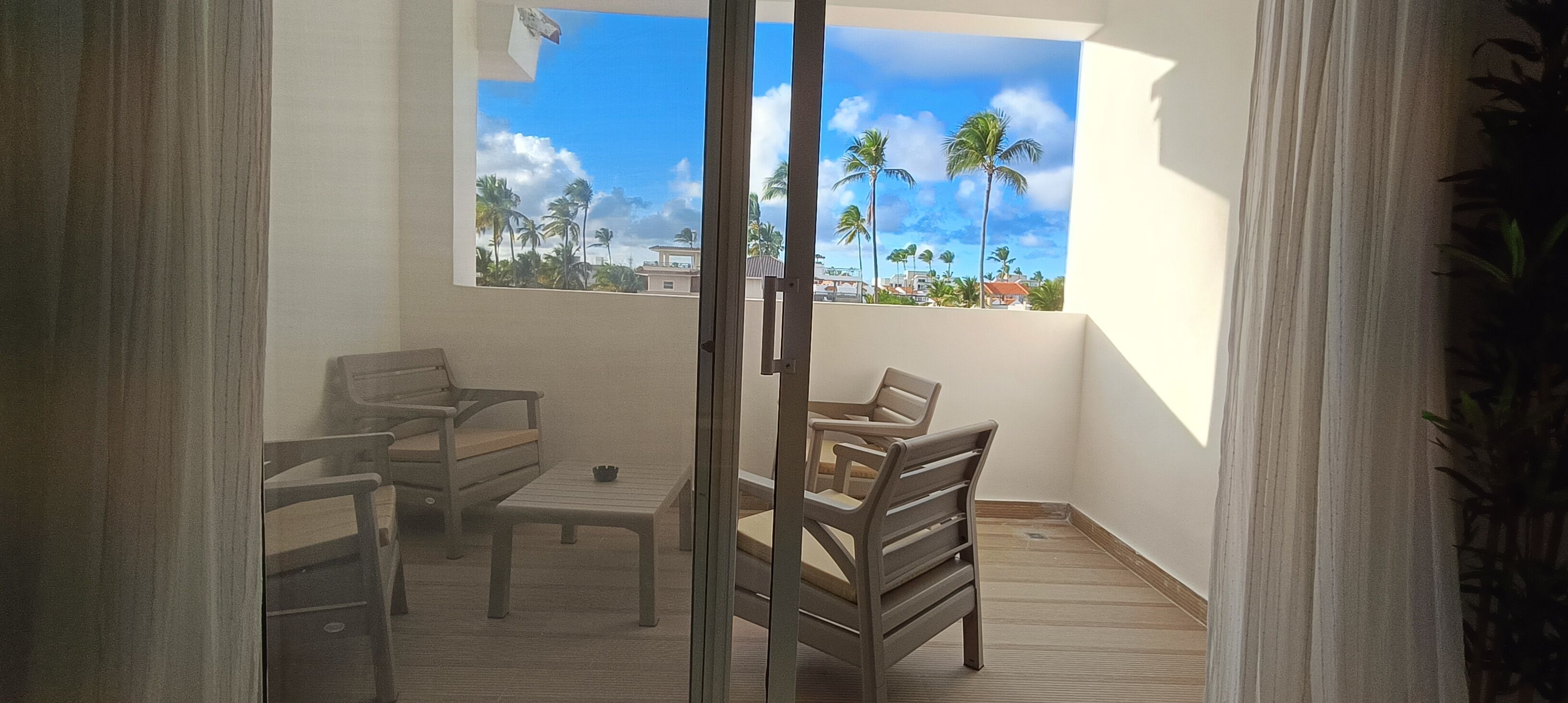 A balcony with a view of palm trees and buildings, wooden furniture, and a window.