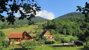 Un paysage rural avec deux maisons traditionnelles en bois, entourées d’une végétation luxuriante et de collines.