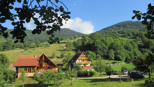 Ländliche Gegend mit zwei traditionellen Holzhäusern, umgeben von üppiger Vegetation und Hügeln.