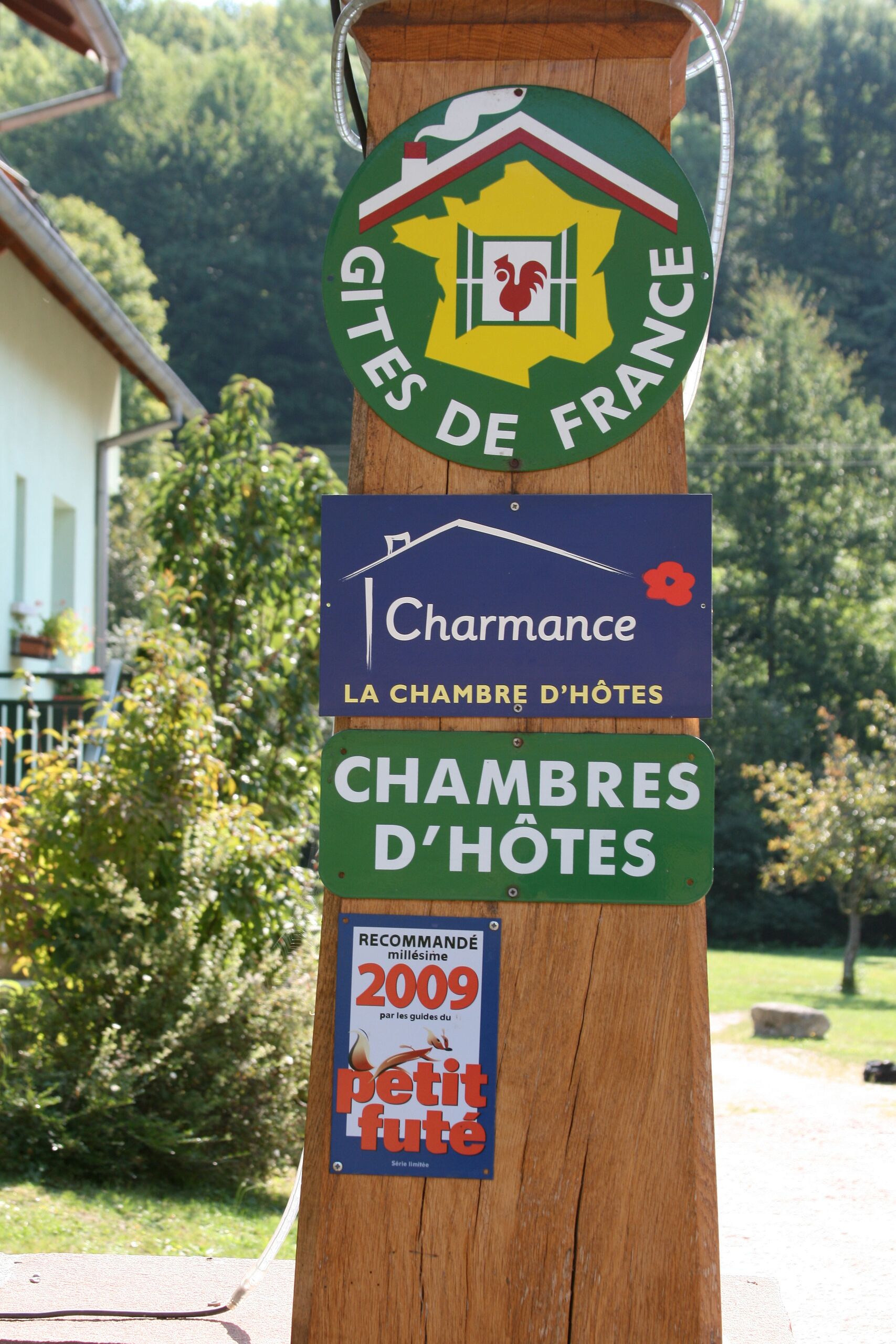 Un poste de madera con varios carteles, incluyendo un logotipo de Gites de France, un cartel para Charmance La Chambre d'Hotes y un letrero de recomendación de 2009.