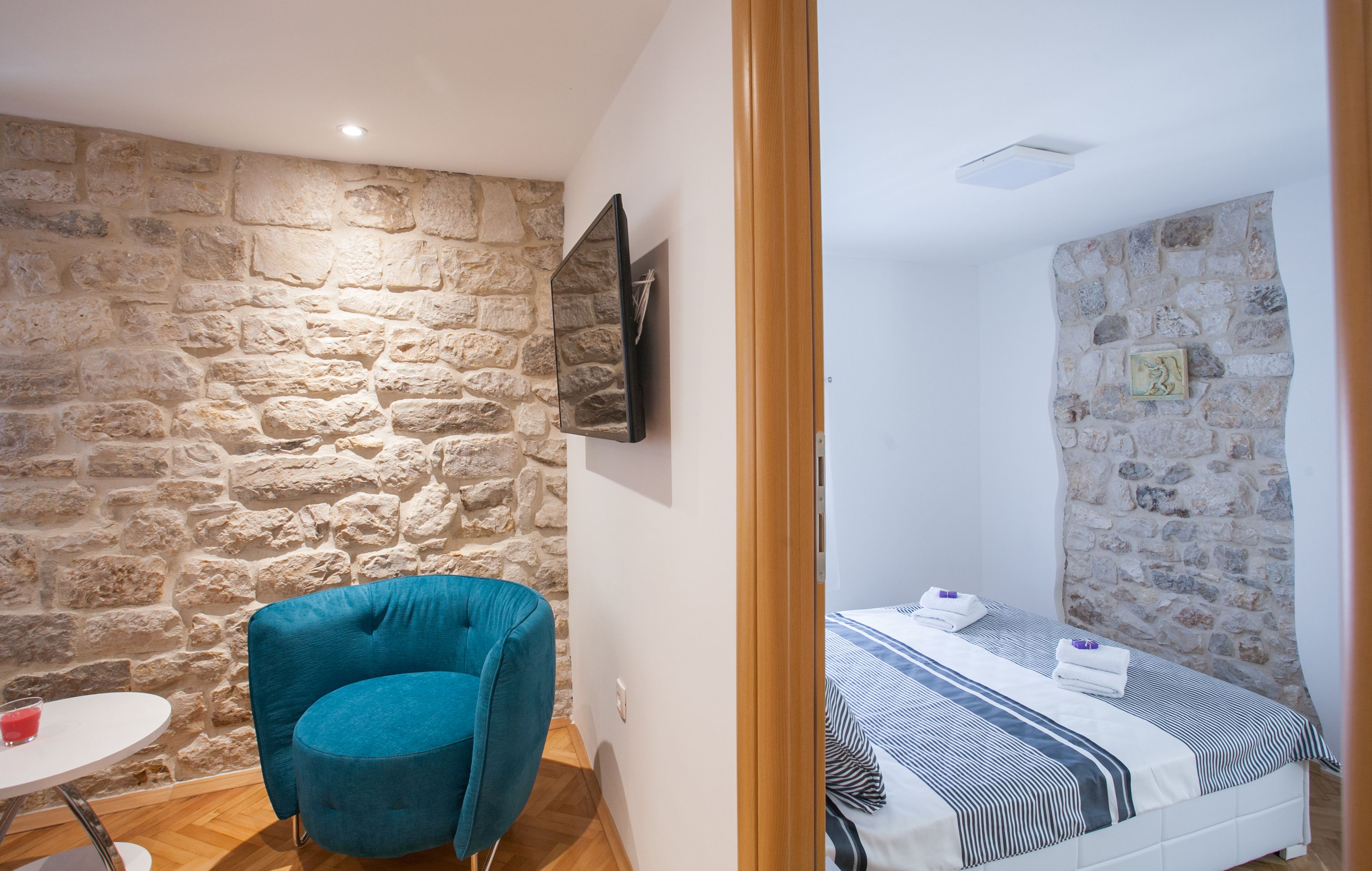 Una habitación con pared de piedra, una cama con ropa de cama a rayas, una silla azul y una mesita.