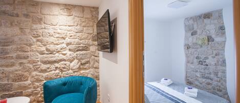 Una habitación con pared de piedra, una cama con ropa de cama a rayas, una silla azul y una mesita.