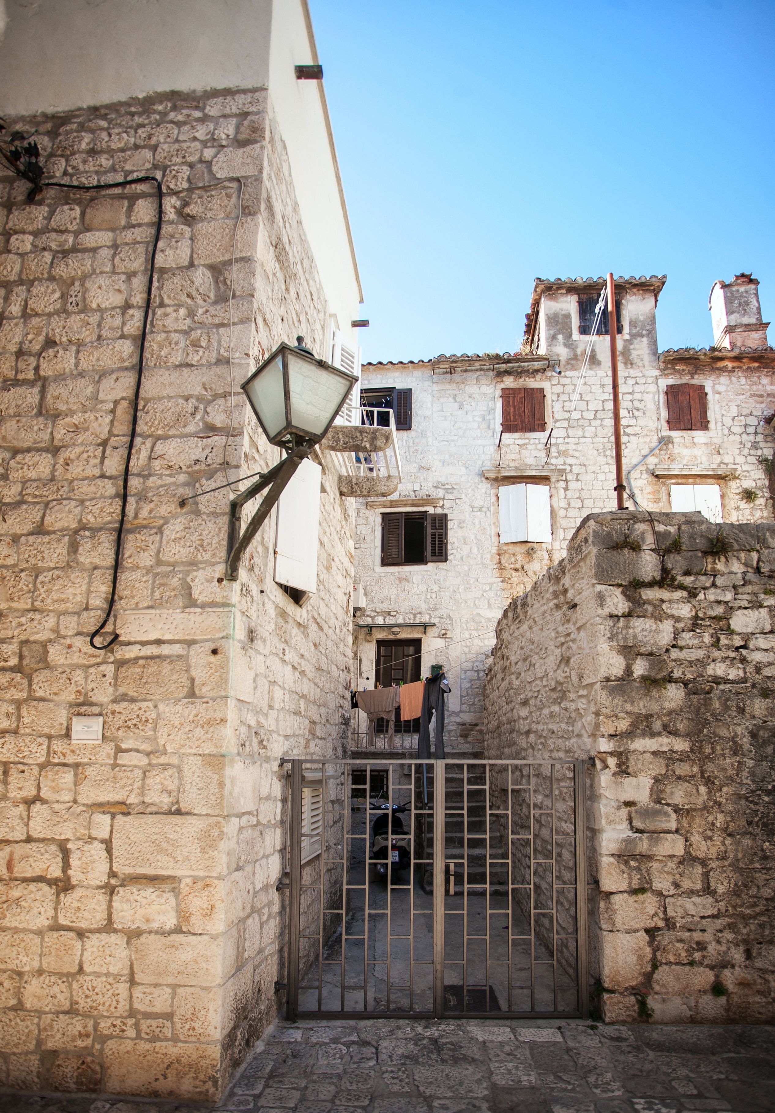 A narrow street with stone buildings, a metal gate, and a hanging light fixture.
