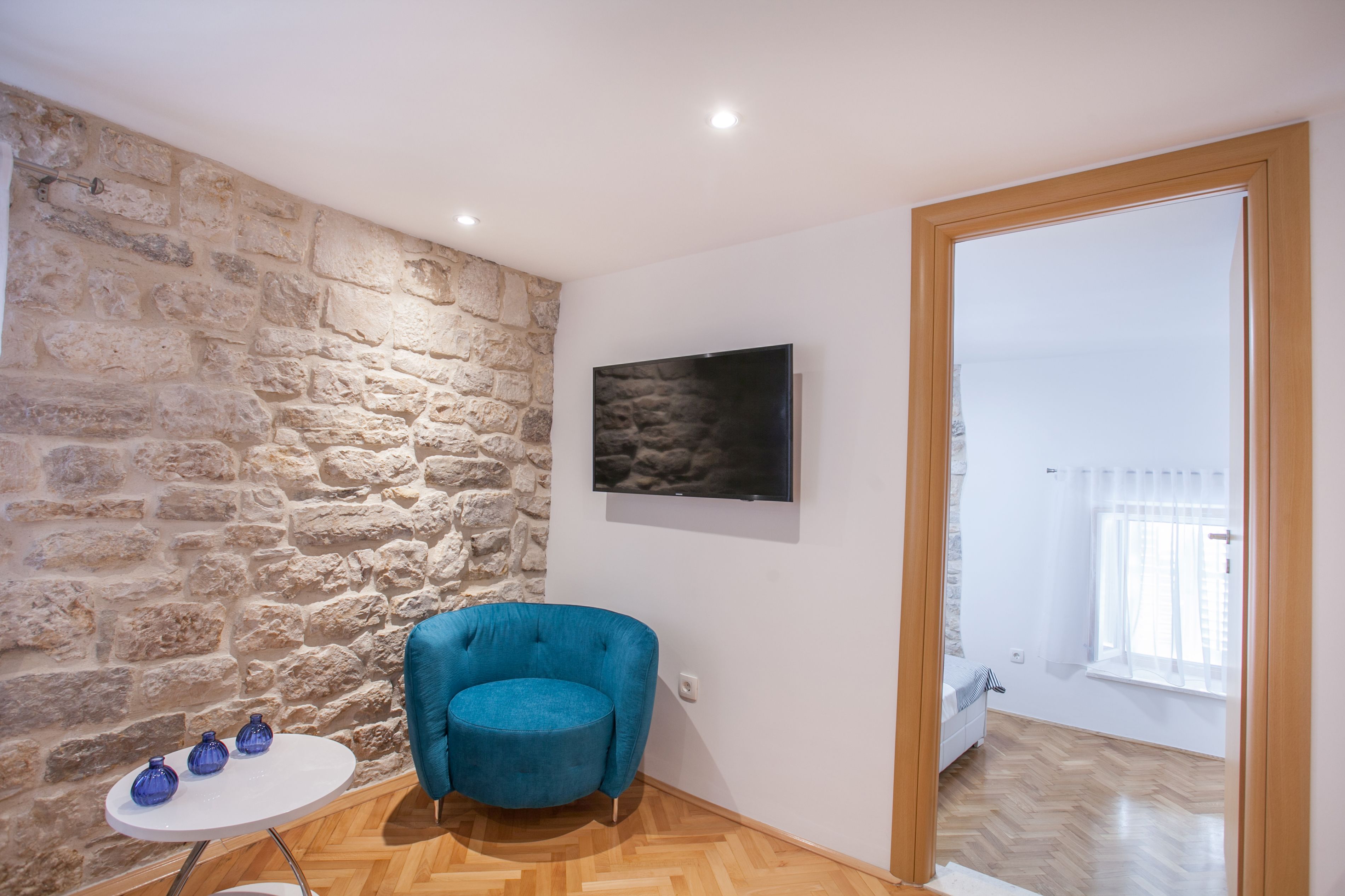 A room with a stone wall, a blue armchair, a small table with vases, a flat-screen TV, and a wooden floor.