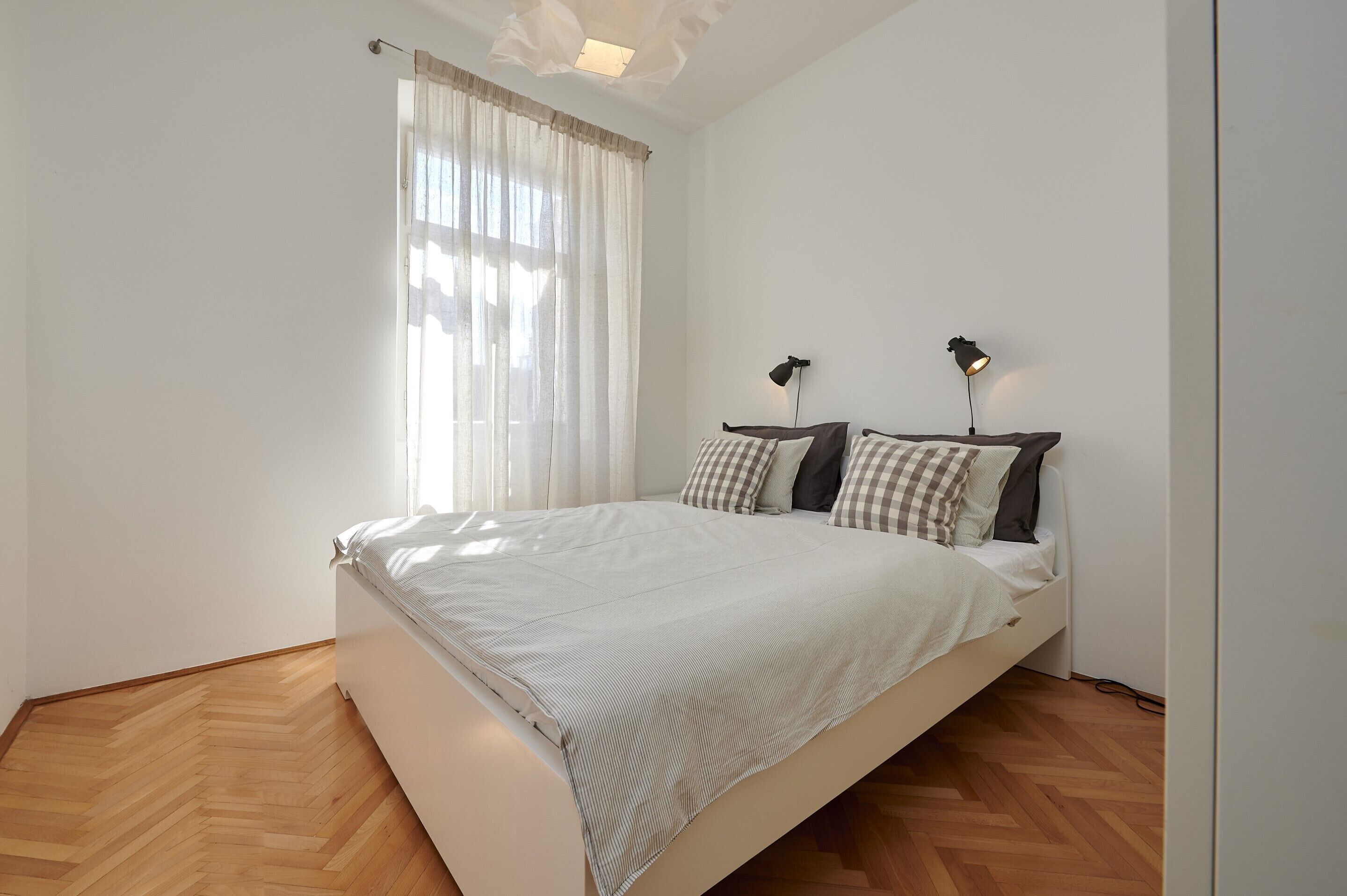 Ein Schlafzimmer mit einem Bett, zwei Nachttischlampen, einem Fenster mit Vorhängen und einem Holzboden.