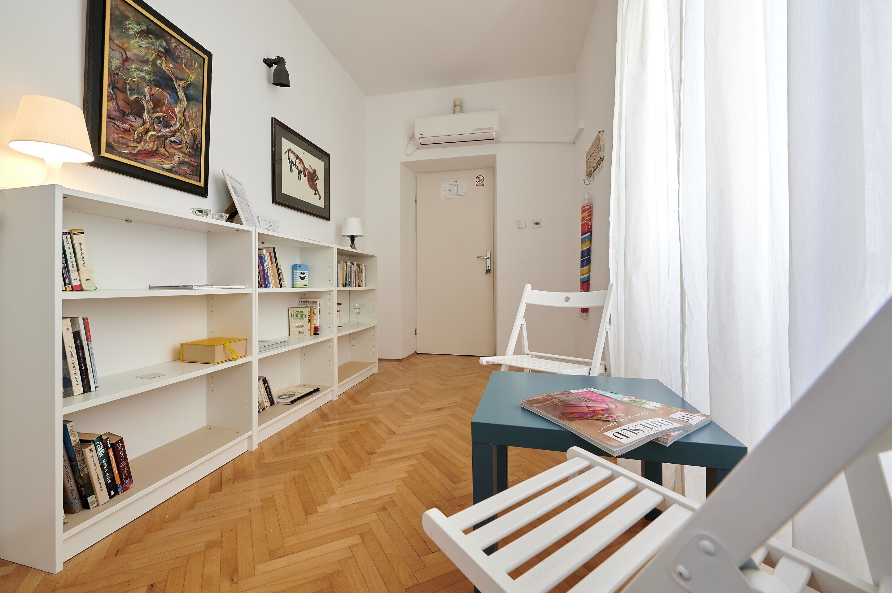 A room with a wooden herringbone floor, a white bookshelf, a table with a magazine, and a white chair.