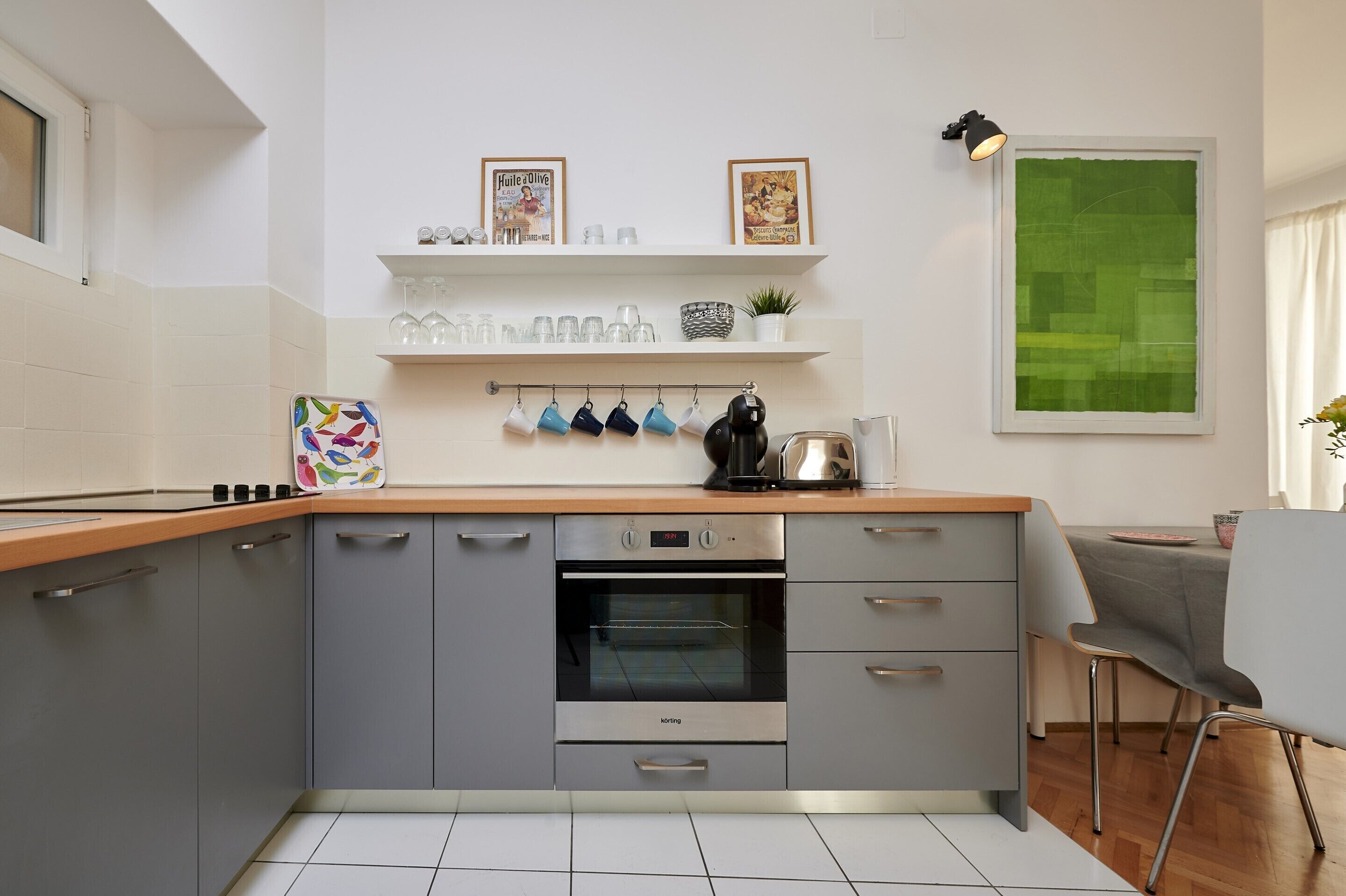 A modern kitchen with grey cabinets, a stainless steel oven, and a wooden countertop. There are shelves above the counter holding various kitchen items and two framed pictures.