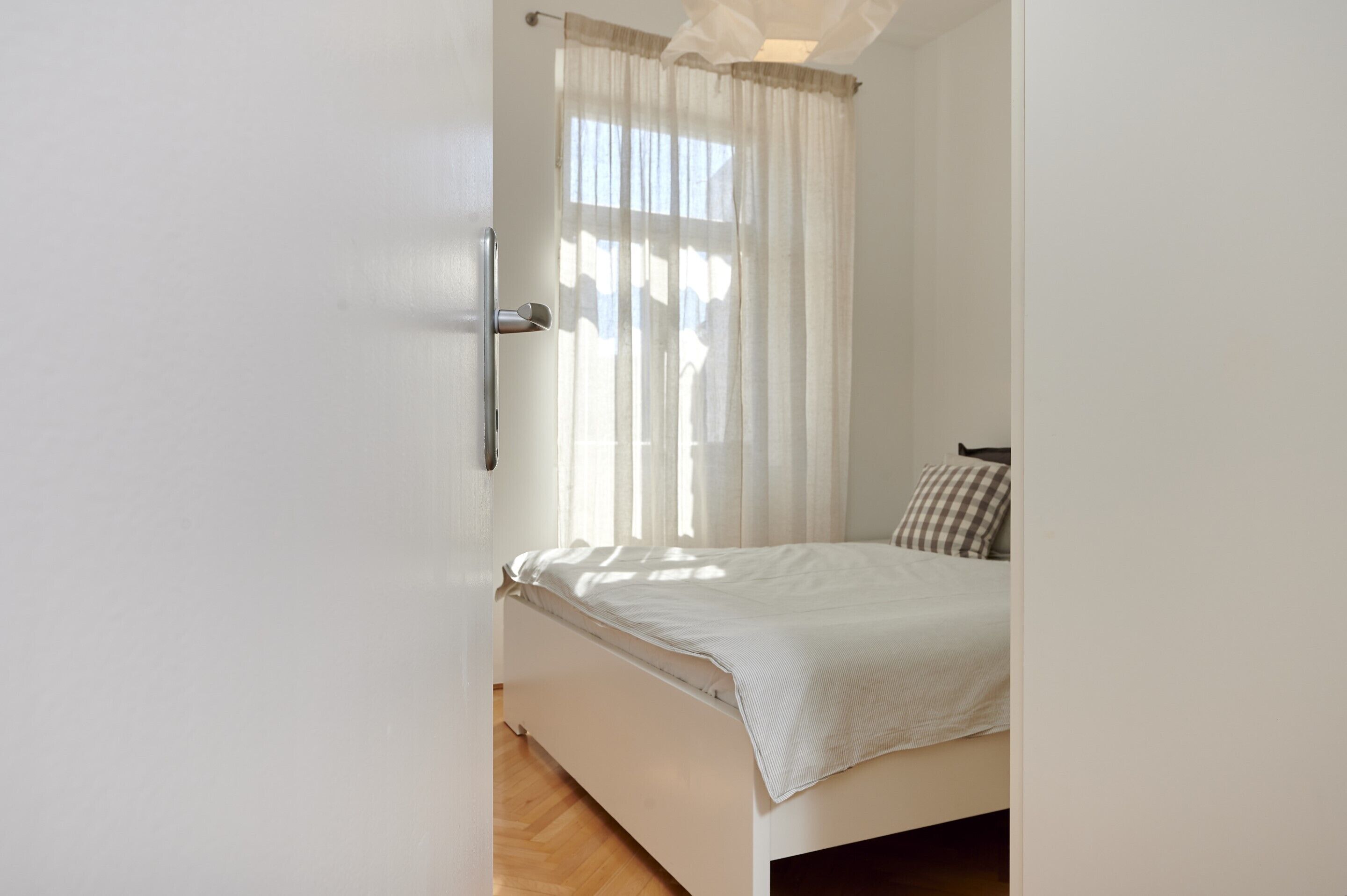 A bedroom with a bed, a window with sheer curtains, and a wooden floor.