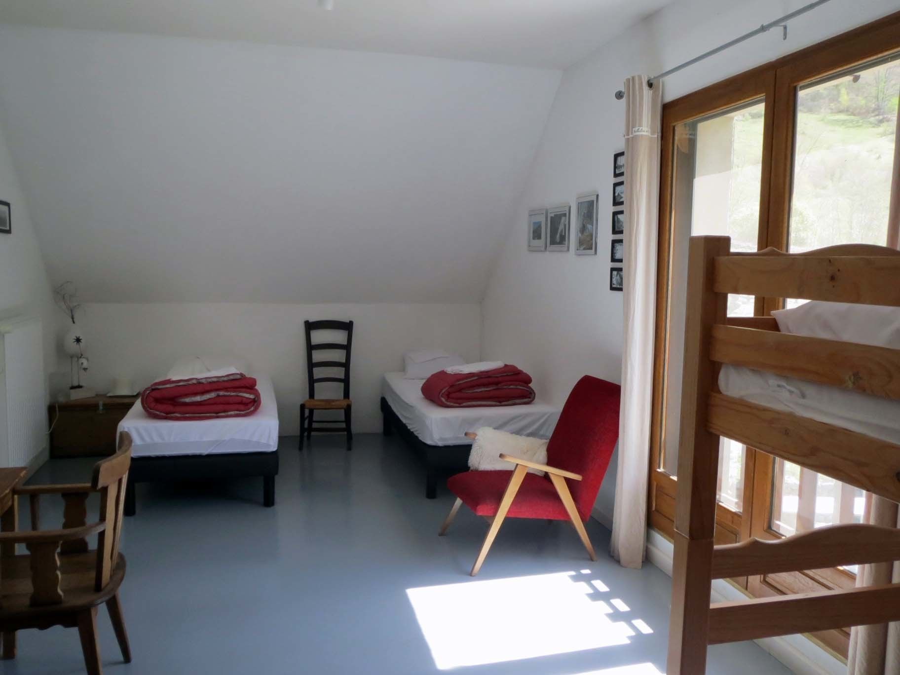 A small, bright room with two single beds, a wooden chair, a desk, and a window.