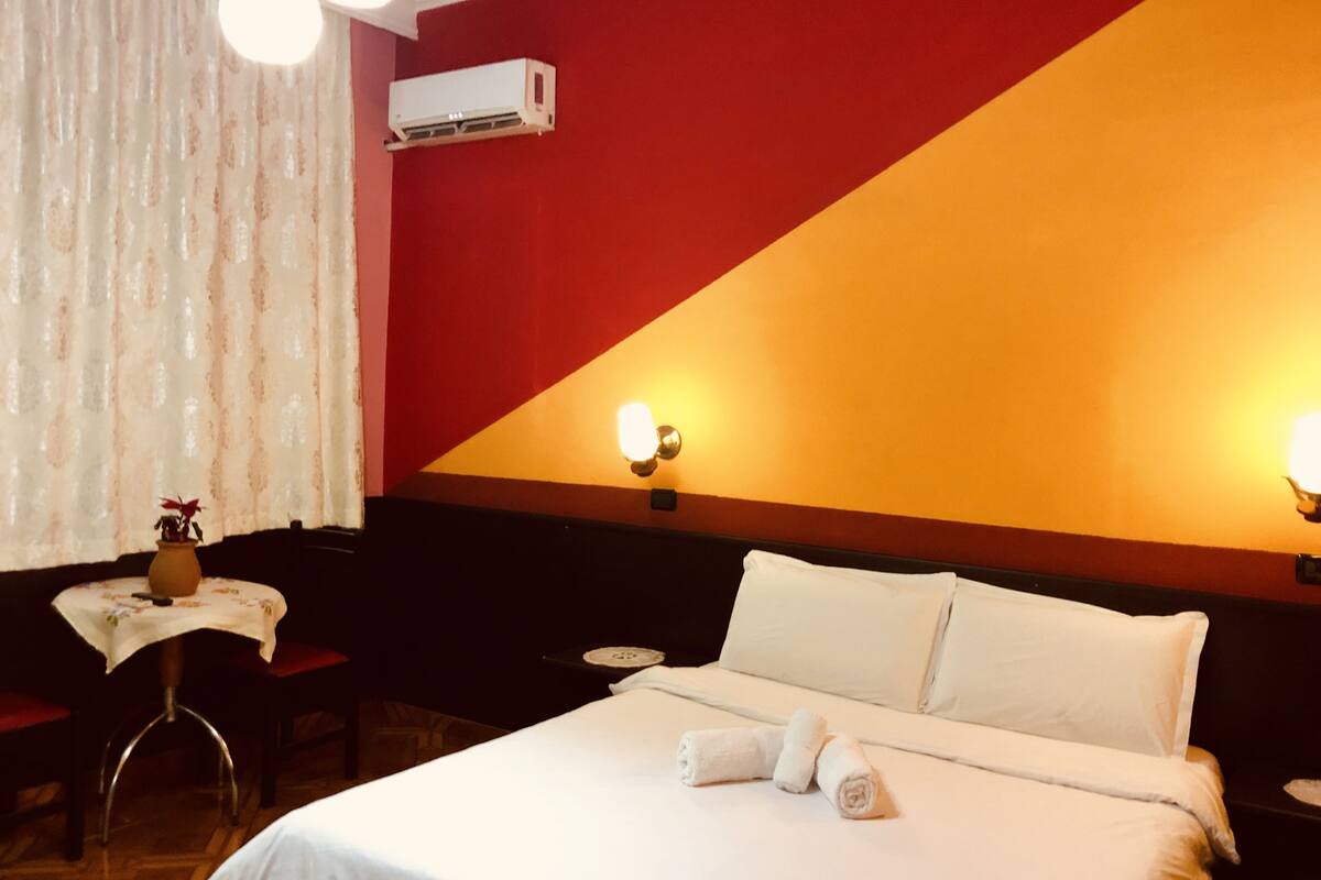 A hotel room with a bed, a small table with a vase, and two wall-mounted lamps.