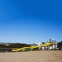Una playa arenosa con un tobogán acuático amarillo y un edificio blanco con detalles azules al fondo.