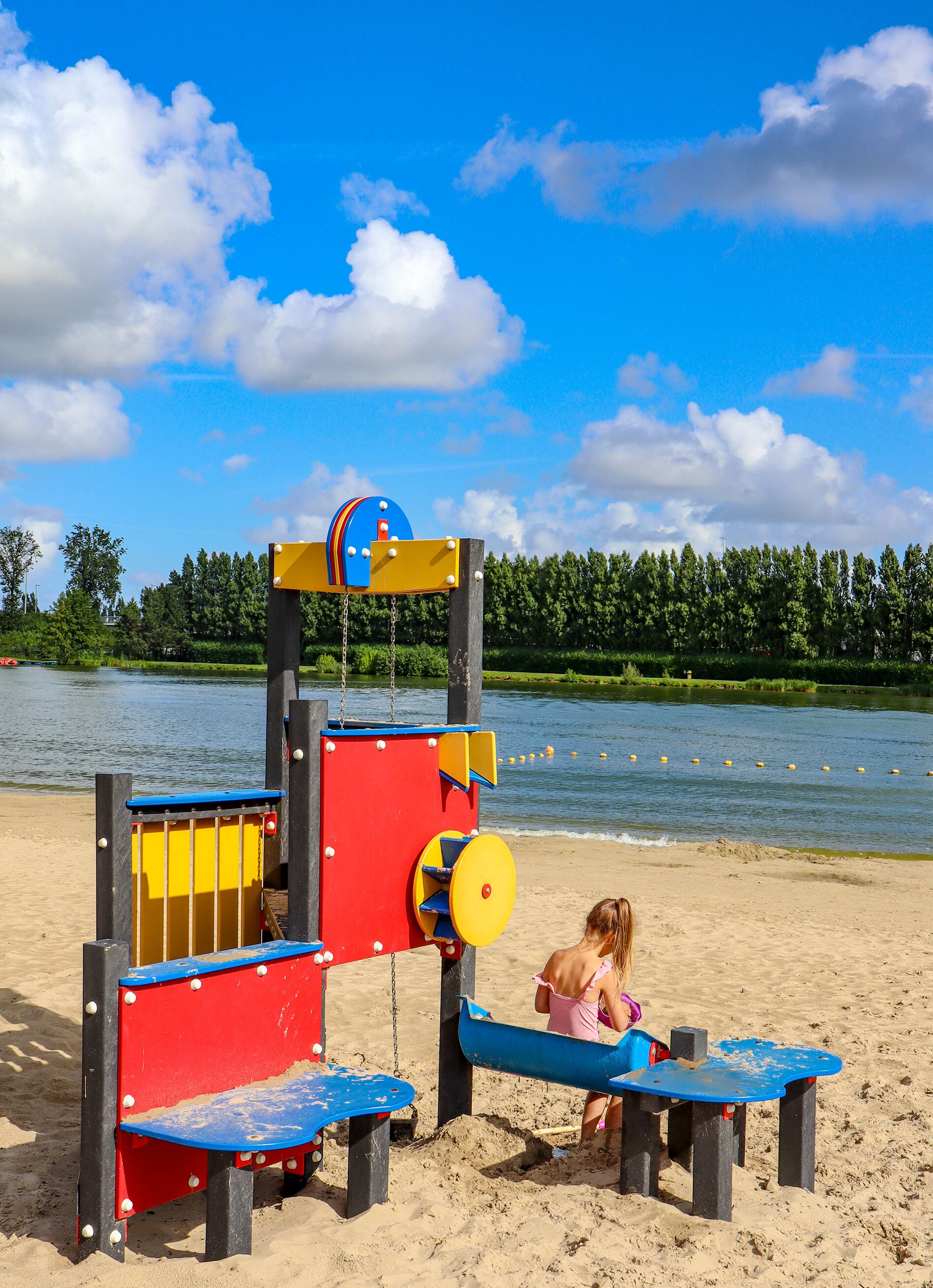 A playground with a slide and climbing structures on a sandy beach near a lake.