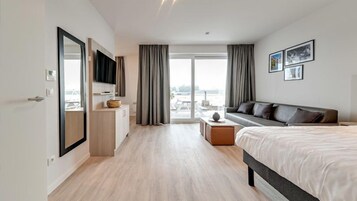 Habitación de hotel moderna con una cama grande, un sofá, un televisor, un escritorio y vistas a la ciudad a través de amplios ventanales.