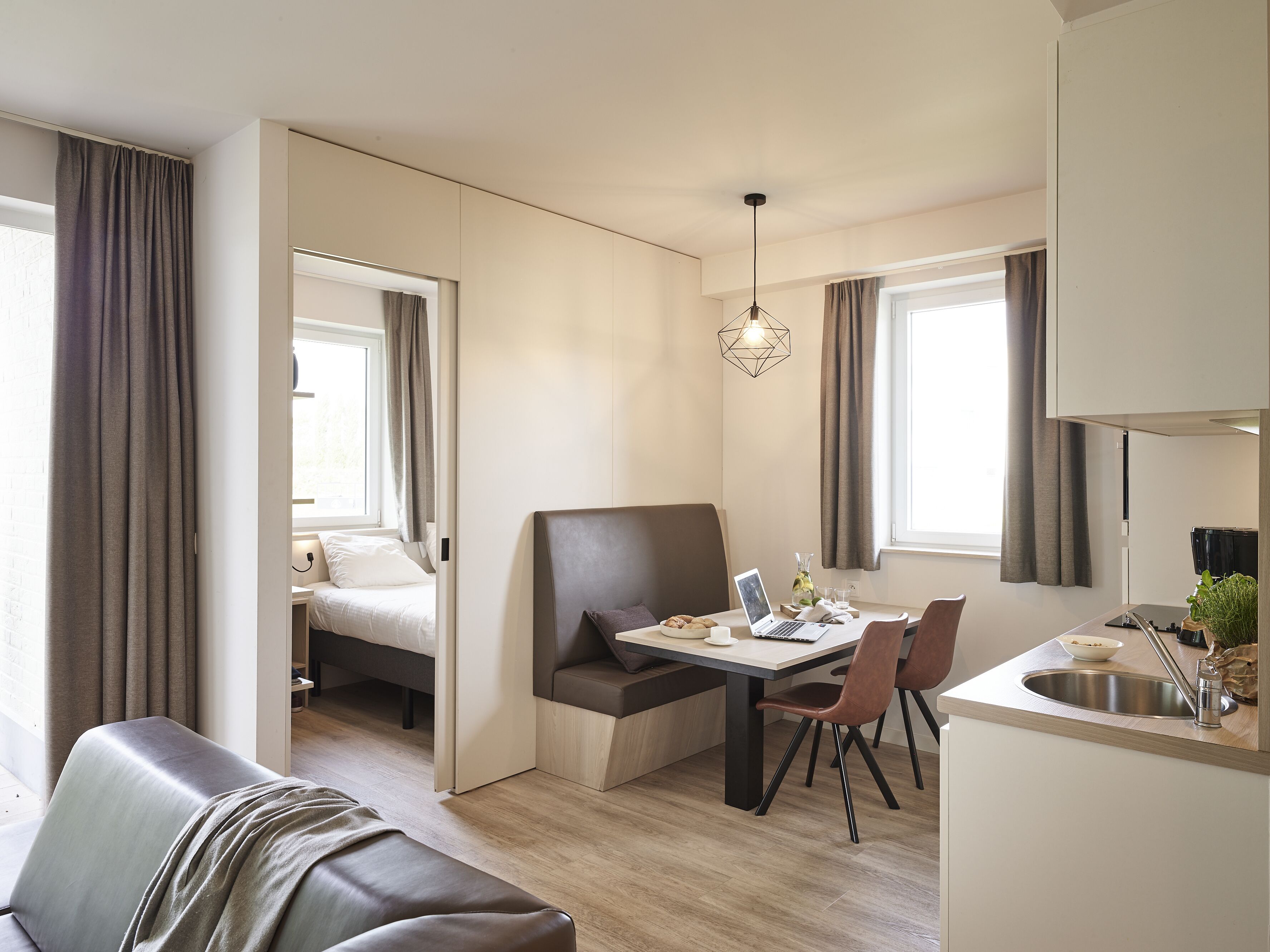 A compact room with a bed, a dining table with chairs, a small kitchenette, and a window with curtains.