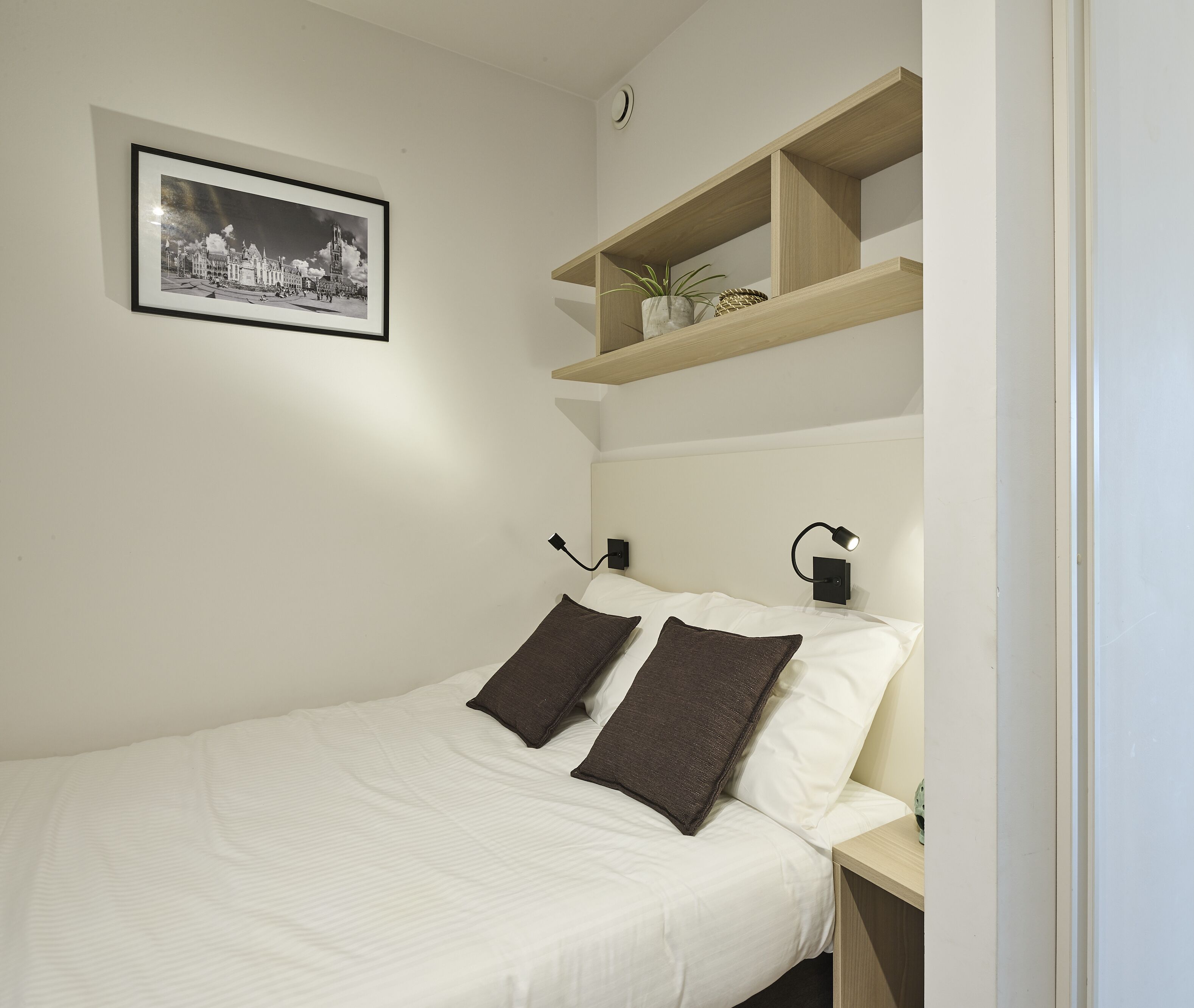 Ein ordentlich bezogenes Bett mit weißer Bettwäsche und zwei dunklen Kissen, eine kleine Ablagefläche mit einer Pflanze und Dekoration, ein gerahmtes Schwarz-Weiß-Foto an der Wand und ein Nachttisch mit einer Lampe.