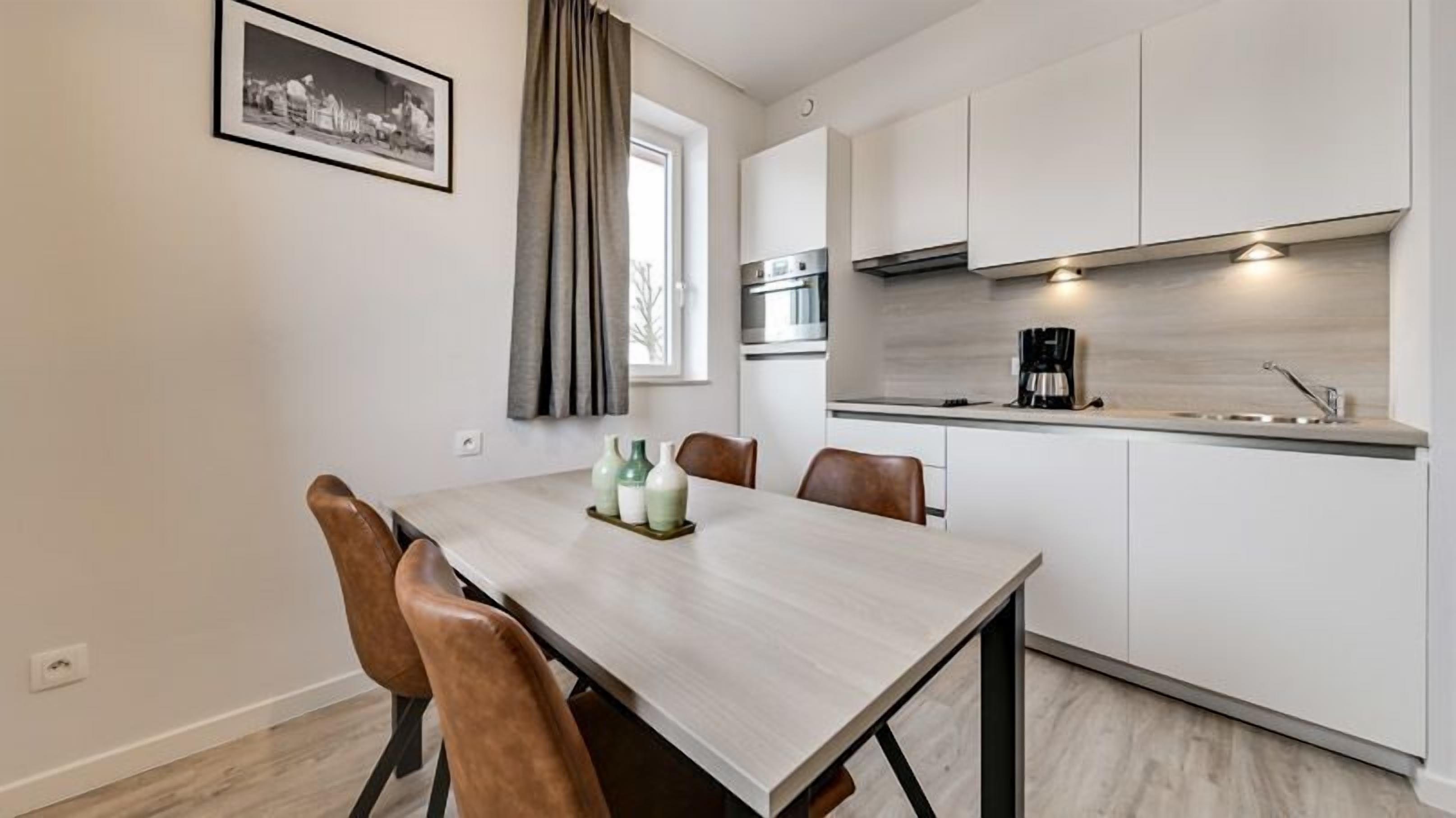 A modern kitchen with white cabinets, a built-in oven, and a coffee maker. There is a dining table with chairs and a window with curtains.