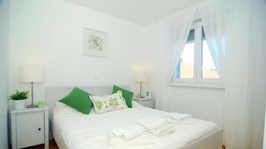 A neatly made bed with white bedding and green pillows, a bedside table with a lamp and a plant, and a window with sheer curtains. - Apartment Flynn (Okrug)