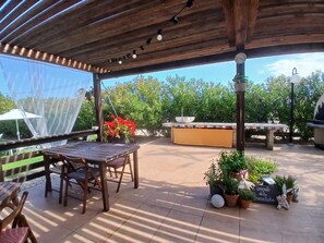 Un patio al aire libre con mesa y sillas de madera, rodeado de plantas en macetas y un área cubierta con mantel amarillo.