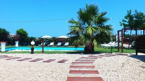 Zona de piscina con sillas reclinables y sombrillas, rodeada de palmeras y un cielo despejado.