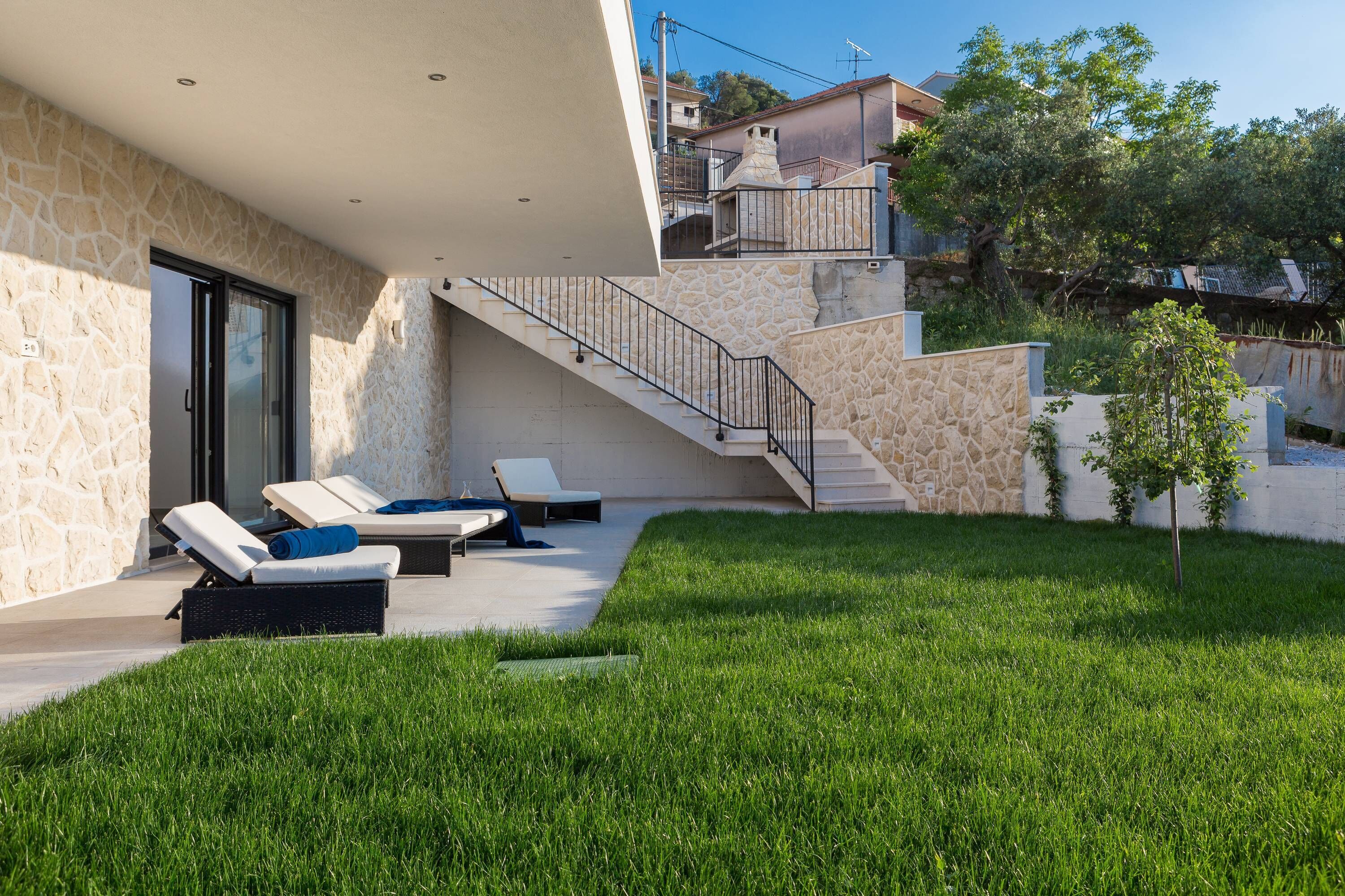 Ein modernes Haus mit Naturstein-Fassade, einer Treppe, die zu einem Balkon führt, und einer Sitzgruppe mit Liegestühlen.