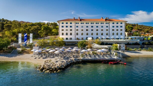 Un grande hotel con facciata bianca e tetto rosso, affacciato su un corpo d'acqua con un molo e ombrelloni.