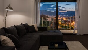 Un soggiorno moderno con un divano scuro, una lampada da terra e vista sulla città al crepuscolo attraverso un'ampia finestra con tende bianche.