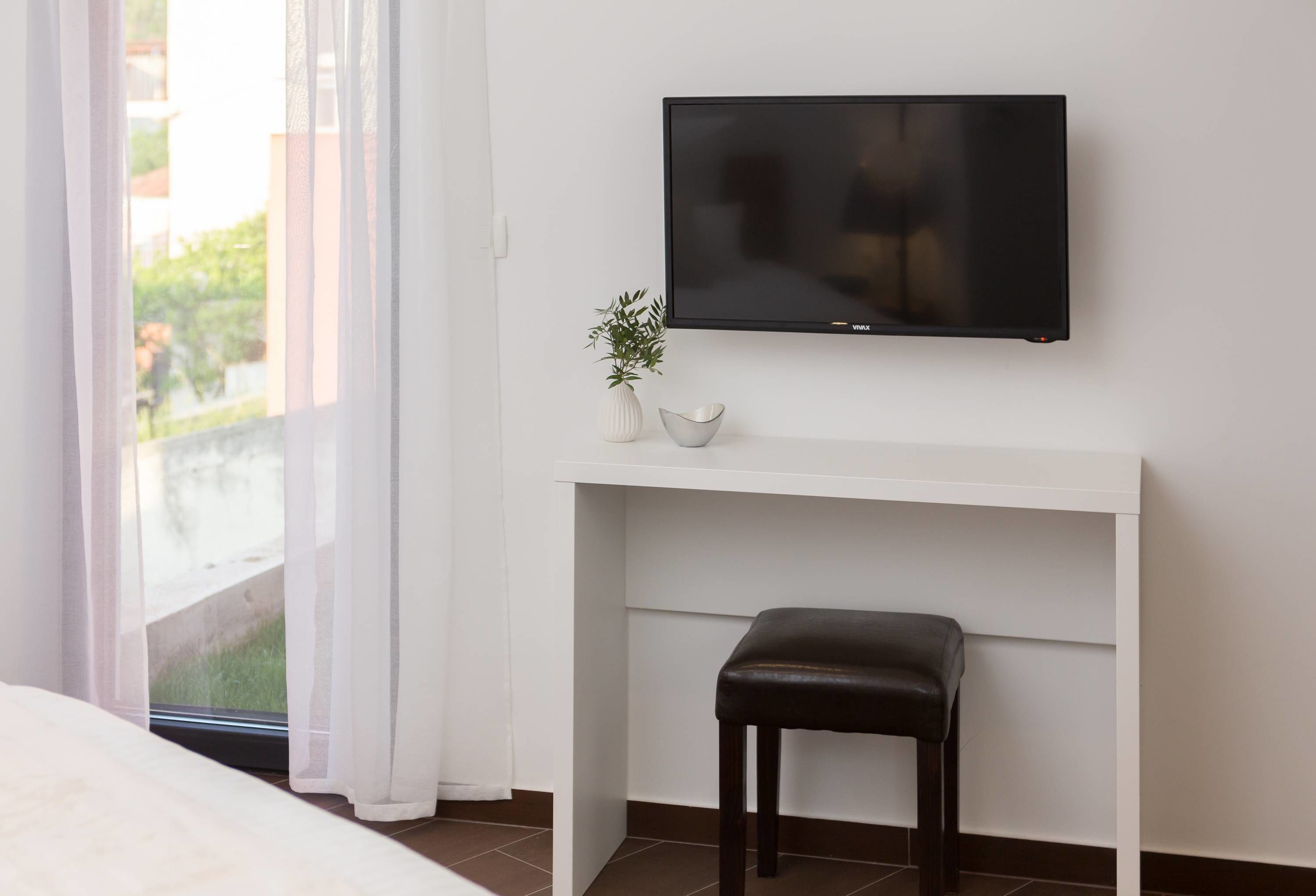A hotel room with a bed, a TV mounted on a white console table, a small plant, and a view of the outdoors through a window with sheer curtains.