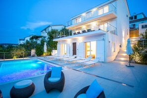 A modern two-story house with a swimming pool, outdoor seating, and a well-lit balcony. - Villa Avalon (Okrug)