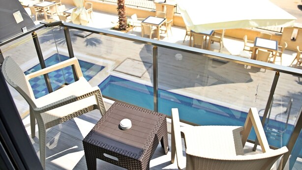 A balcony with wicker furniture, a small table with a bottle, and a view of a pool and outdoor seating area. - Kaptan Suit Otel (Silifke)