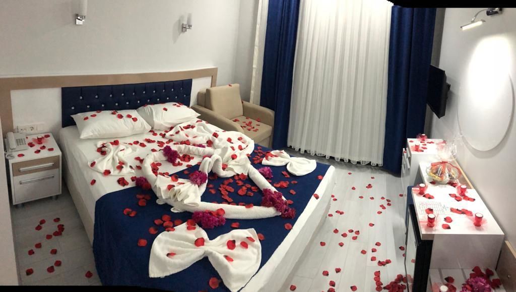 A hotel room with a bed covered in rose petals and a towel with a bow, a bedside table with a bottle, and a chair.